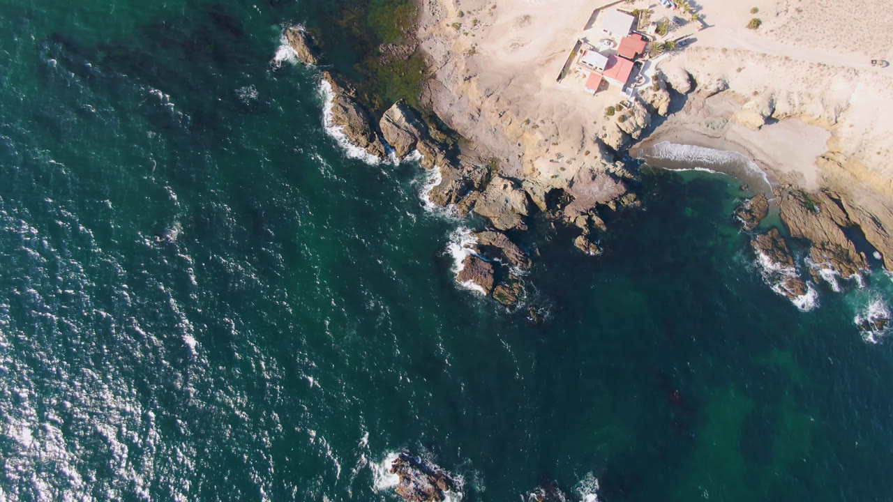 Aerial view of blue-green water, rocky shore, sand, and a coastal house.