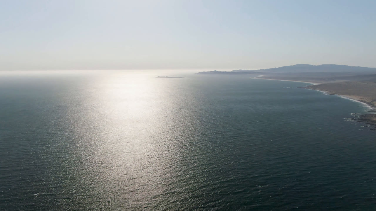 High aerial view over Bahia Ascuncion with sun glare on the ocean, a sandy coastline to the right, distant land on the horizon, and no visible people.