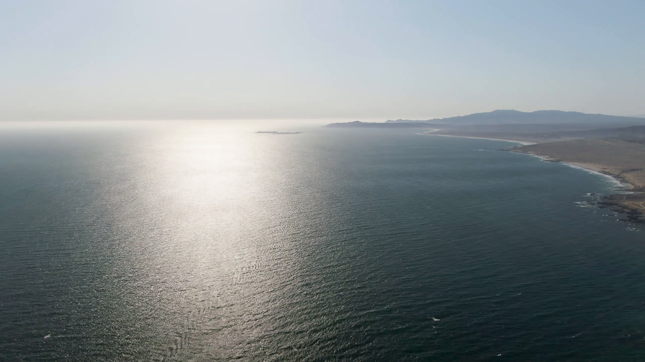 Aerial frame over Bahia Ascuncion showing bright reflected sunlight across the water, a long tan shore on the right, hazy distant hills, and no visible people.
