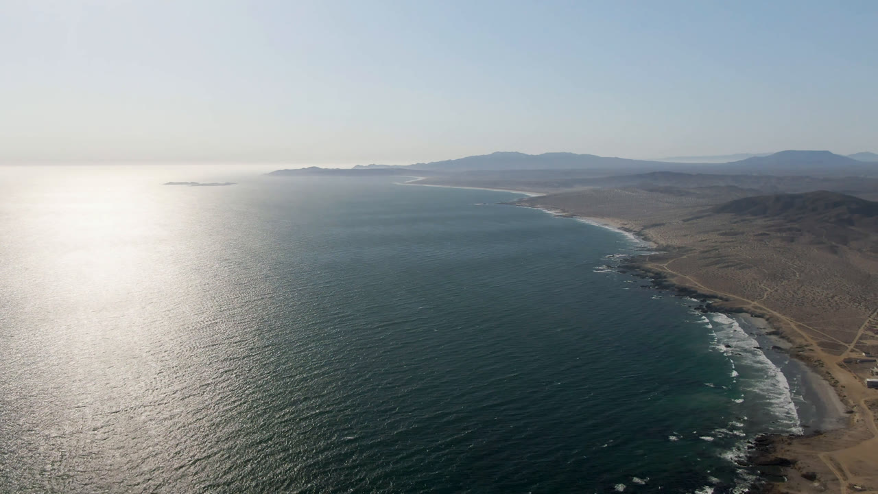 Aerial view over Bahia Ascuncion with bright ocean glare on the left, a sandy shoreline on the right, dry coastal hills beyond, distant mountains, and no visible people.