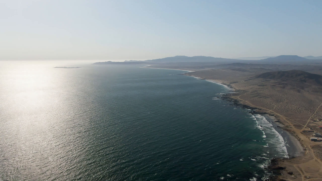 Drone view of Bahia Ascuncion coastline with dark blue water, a curved sandy beach, white surf bands, sparse dry terrain, distant mountains, and no visible people.