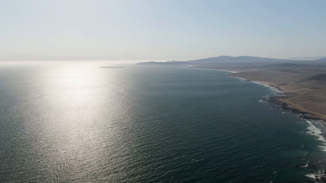 Opening aerial frame over Bahia Ascuncion showing bright sun reflection on blue-green ocean, a curving sandy shoreline, low brown land, distant mountains, and no visible people.