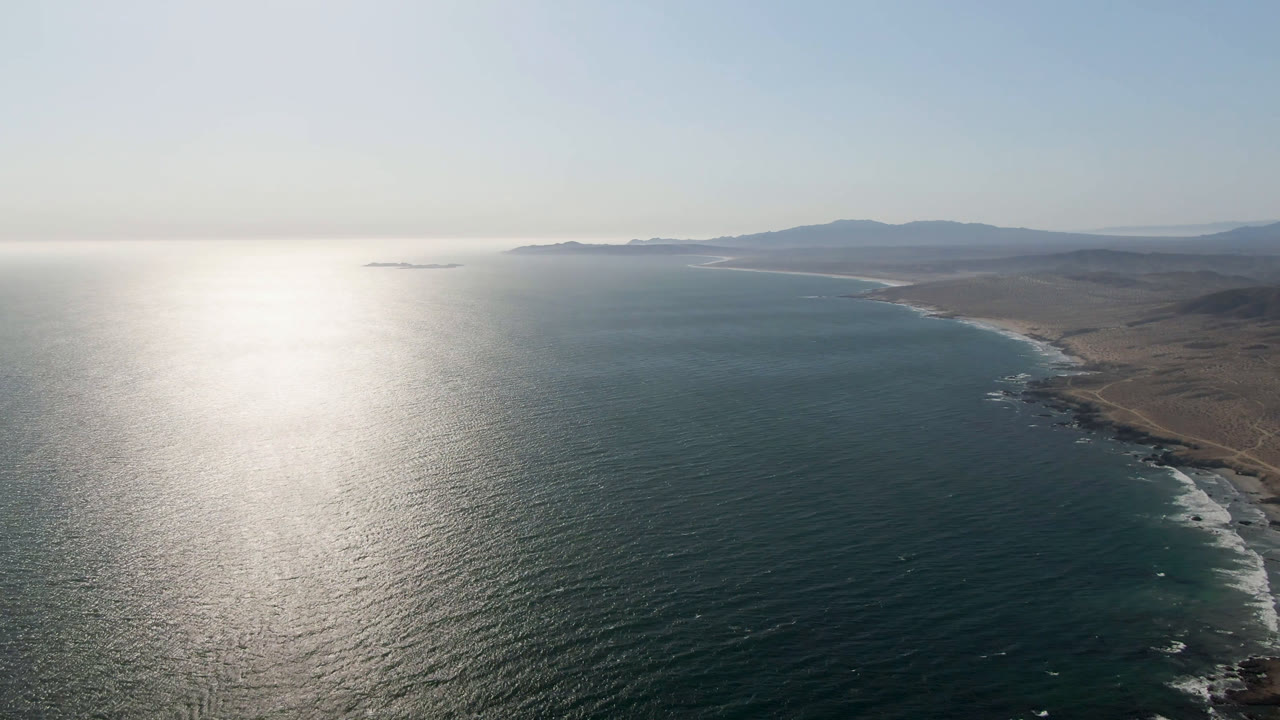 Aerial view over Bahia Ascuncion with the sun glare centered on the water, the coastline bending along the right side, hazy mountains beyond, and no visible people.