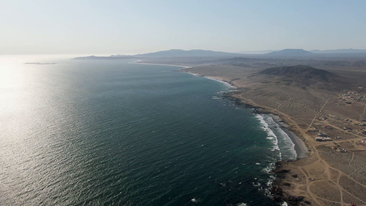 Aerial coastline with ocean, surf, dry land, and distant hills.