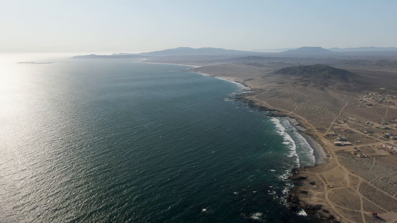 Aerial frame from Bahia Ascuncion showing the bay curving into the distance, white surf along the shore, tan dirt roads across the coastal plain, low hills beyond, and no visible people.