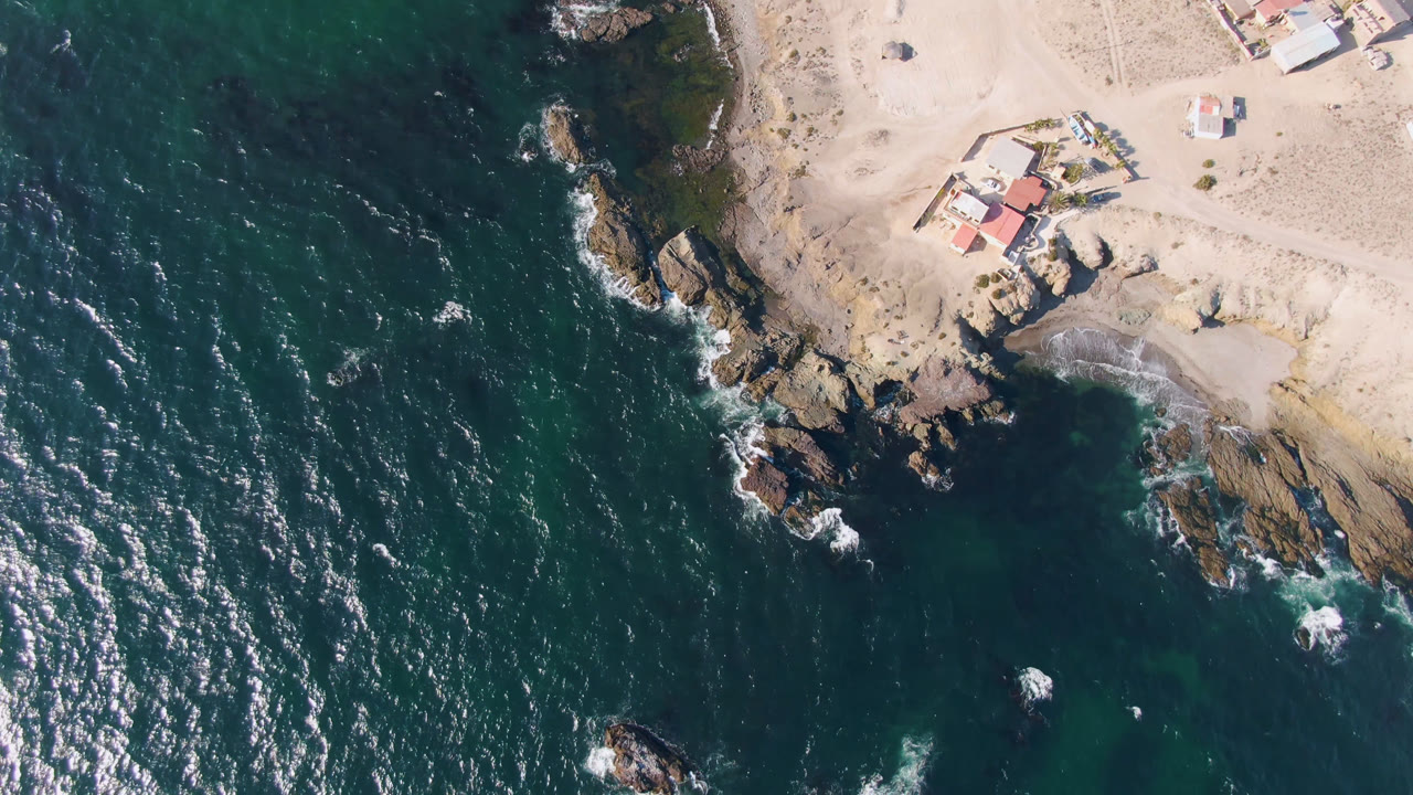 Overhead view of Bahia Ascuncion coastline with dark teal water on the left, tan sand at the top, rocky shoreline in the center, small coastal buildings, and no visible people.
