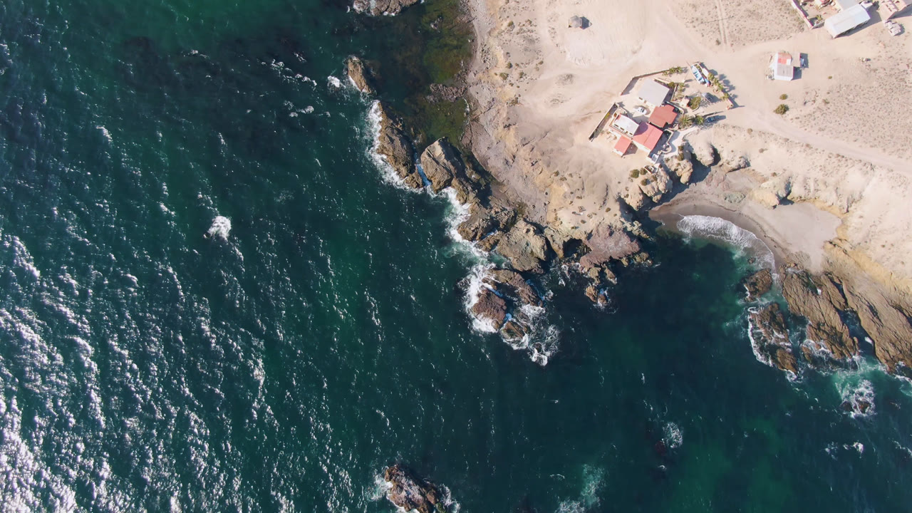 Overhead coastline with teal ocean, rocks, sand, and a small red-roofed structure.
