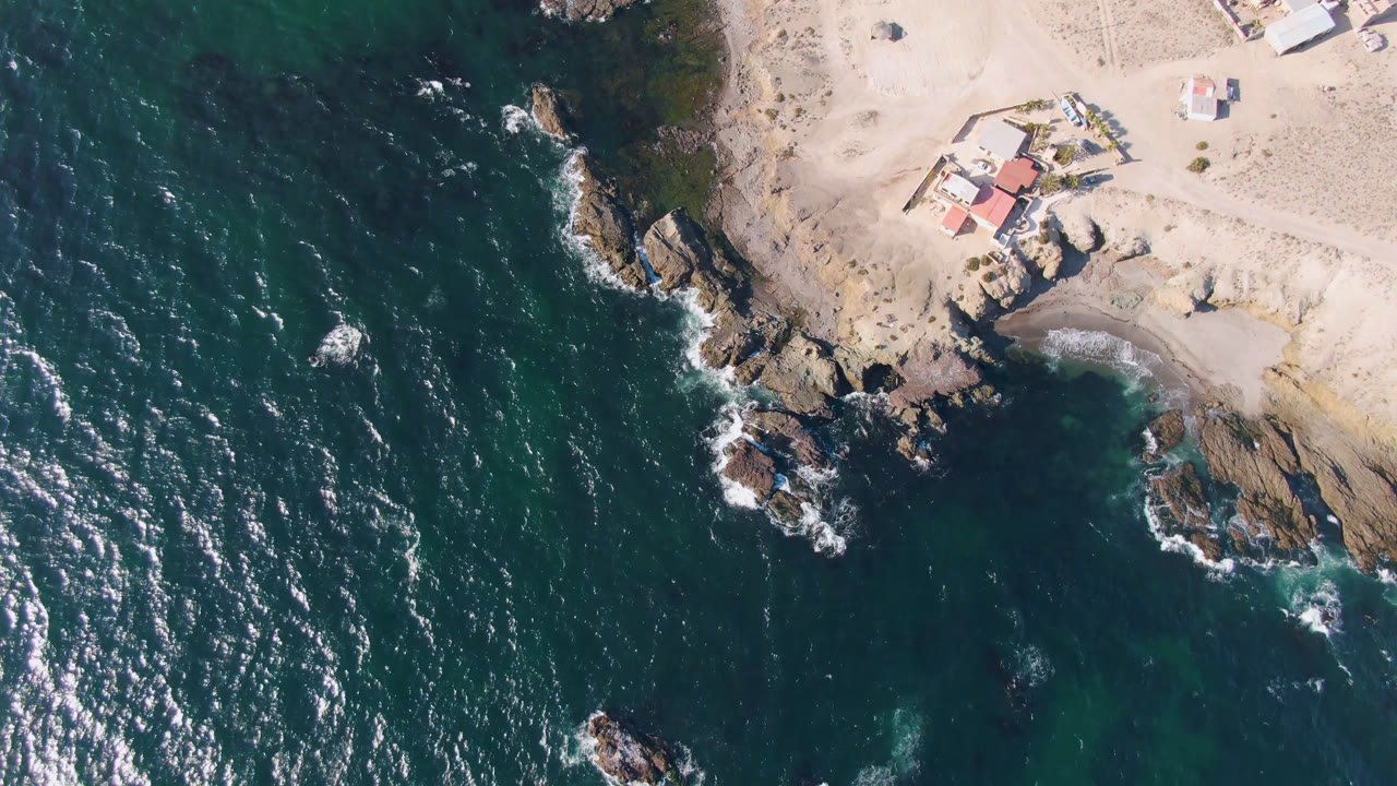 At 0:01, the Bahia Ascuncion shoreline remains centered, with brighter surf flecks across the water, pale sand at the top, coastal rocks, small structures, and no visible people.