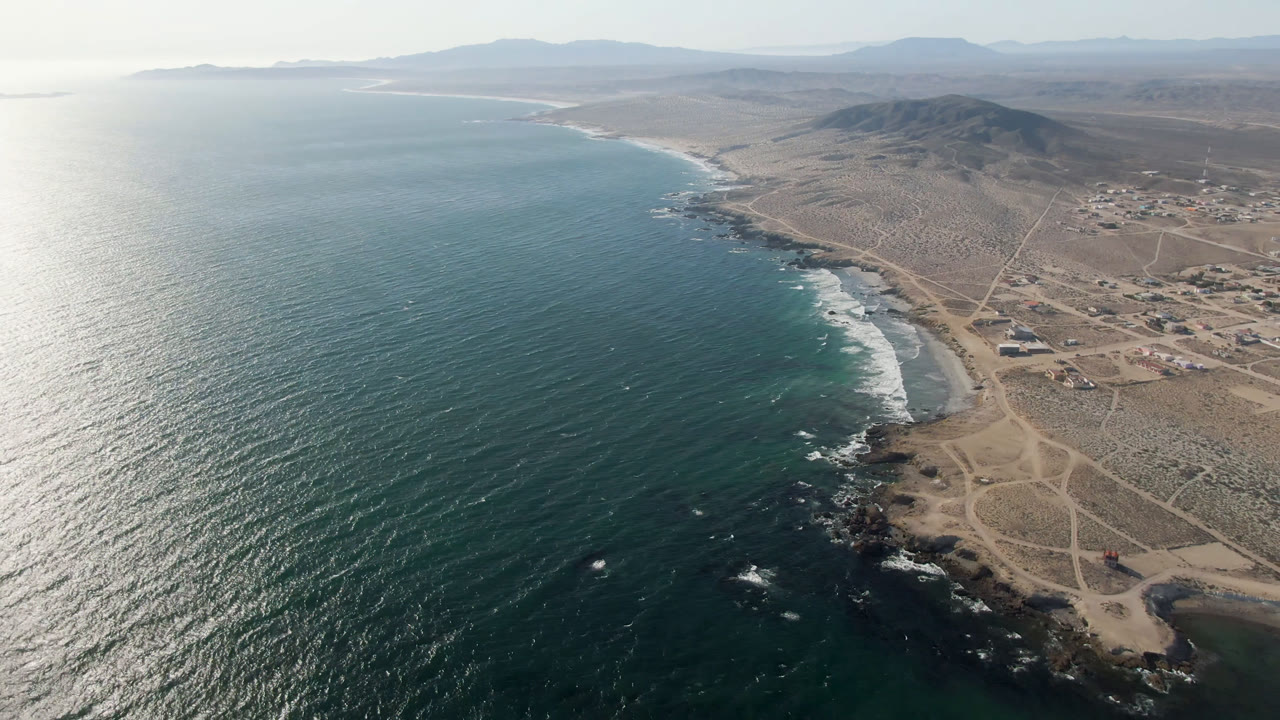 High aerial view over Bahia Ascuncion with teal ocean at left, a rocky point and dirt tracks below, scattered buildings inland, distant hills, and no visible people.