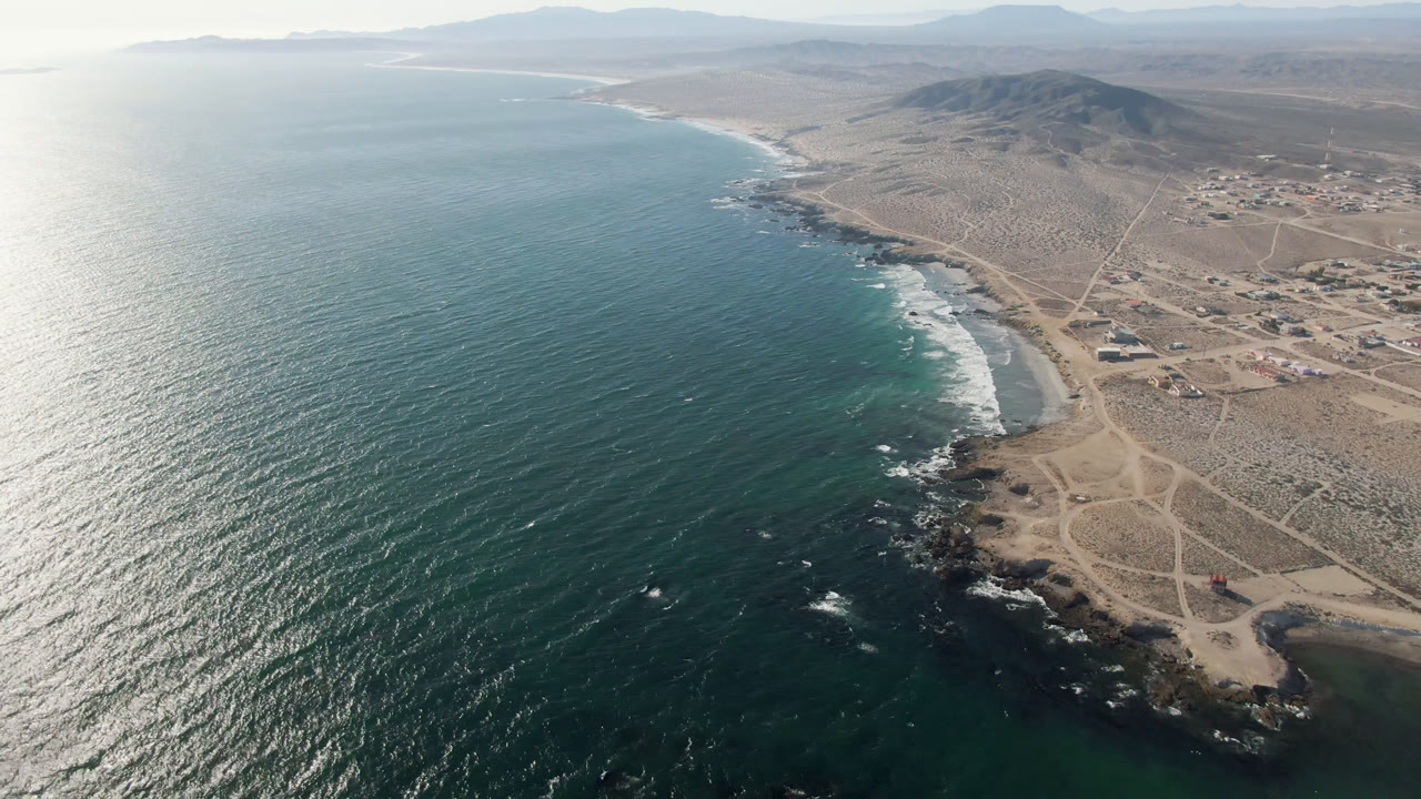 Aerial frame of Bahia Ascuncion showing bright sun reflection on the ocean, white surf along the curved shore, a rocky headland, inland roads, low buildings, and no visible people.