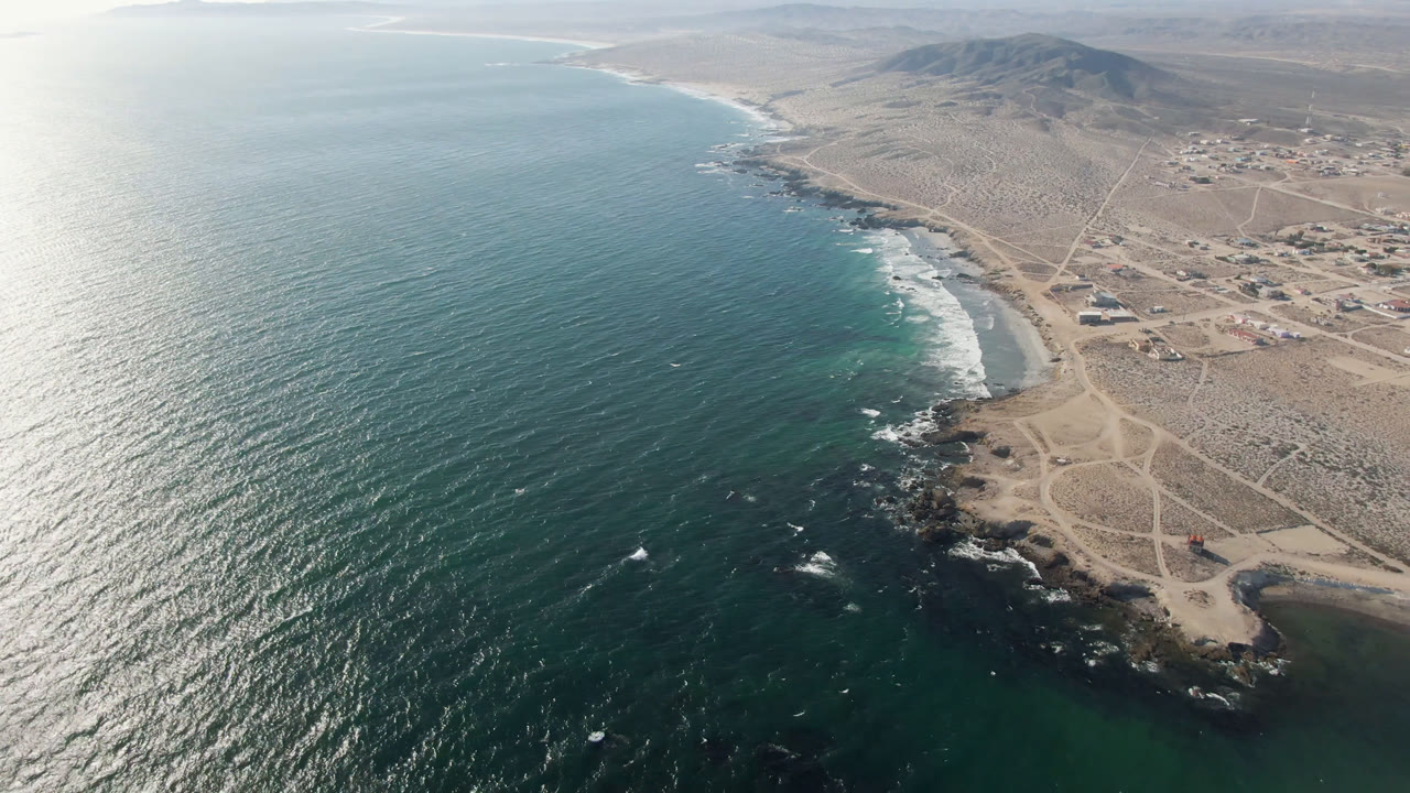 Wide drone view of bay water, shoreline surf, rocky coast, roads, and inland settlement