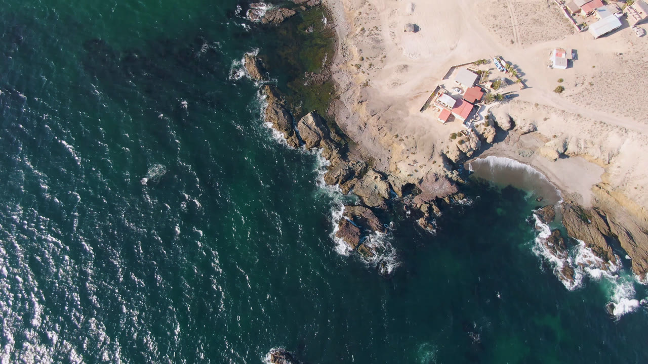 Aerial frame over Bahia Ascuncion showing white surf around shoreline rocks, deep green-blue water, pale beach sand, and roofed houses inland, with no visible people.
