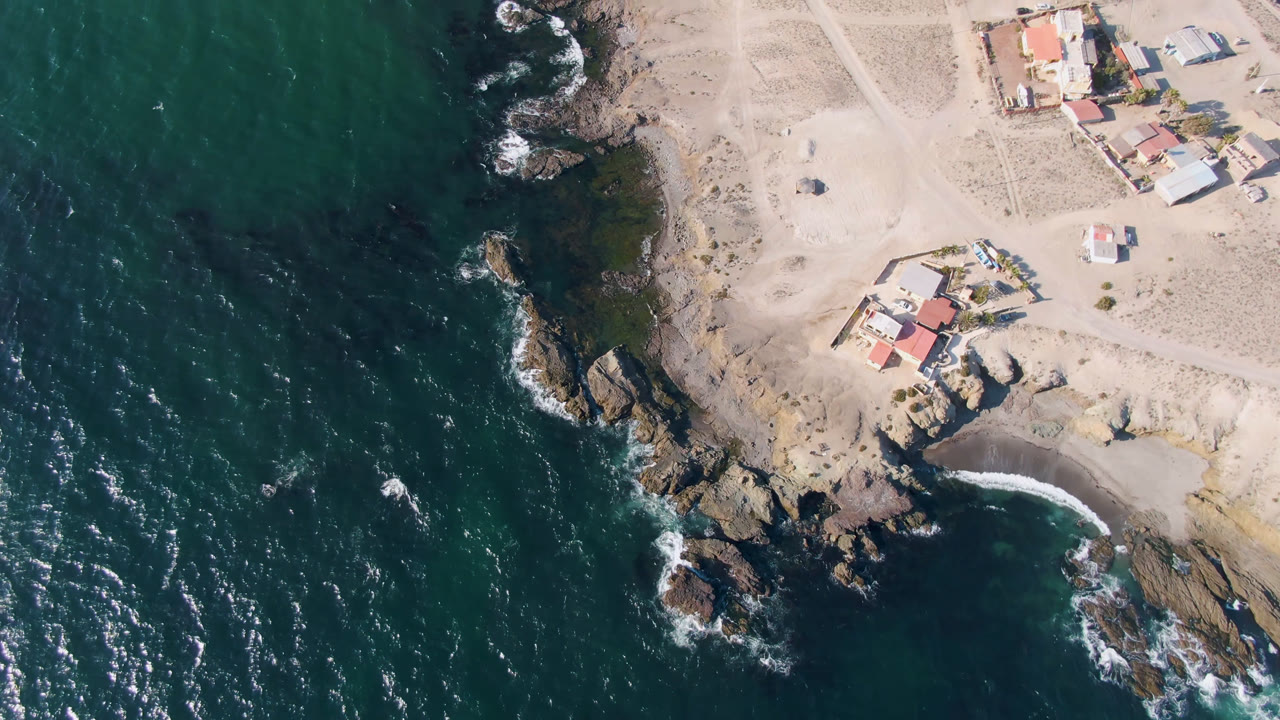 Overhead frame of Bahia Ascuncion coastline with dark teal water on the left, rocks along the shore, sandy ground, several small houses, and no visible people.