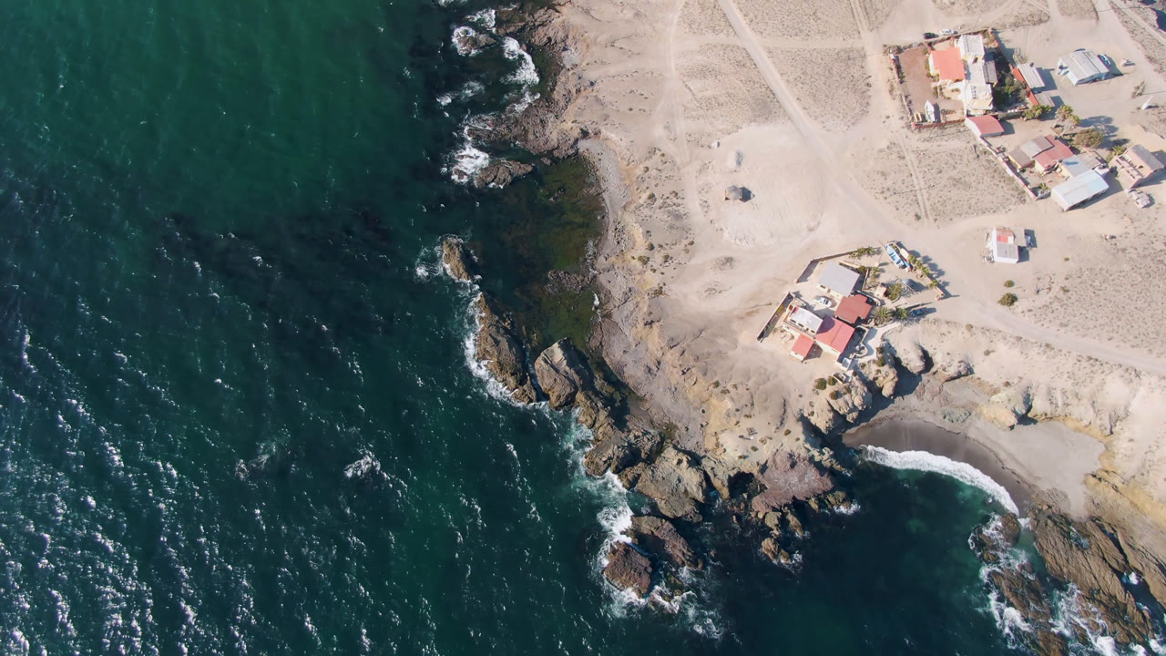 Aerial view over Bahia Ascuncion with ocean occupying the left side, surf breaking on rocks, pale sand roads inland, clustered houses near the upper right, and no visible people.