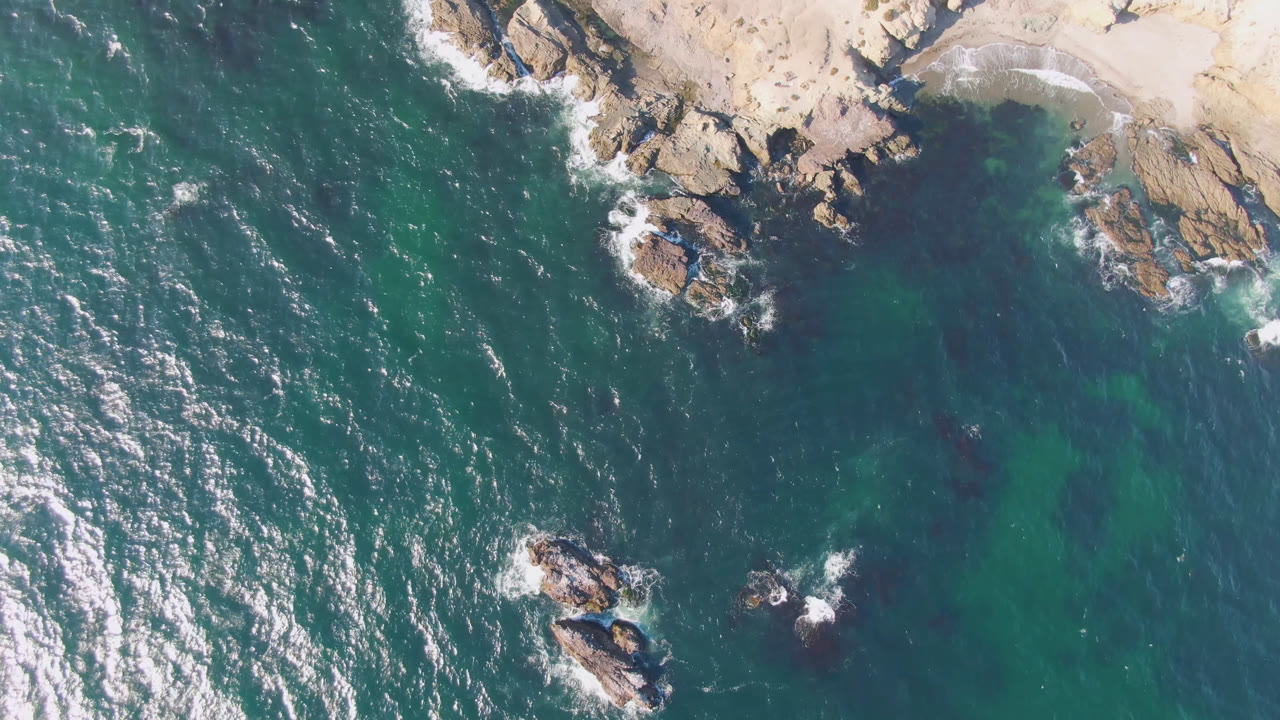 Overhead frame from Bahia Ascuncion showing teal ocean beside tan rocks and a pale sandy beach at the top edge, with no visible people.
