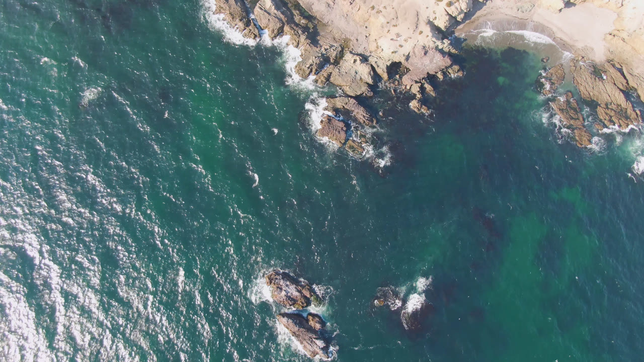 Aerial frame from Bahia Ascuncion with sunlit ripples across teal water, rocky shore along the top, and small offshore rocks below, with no visible people.