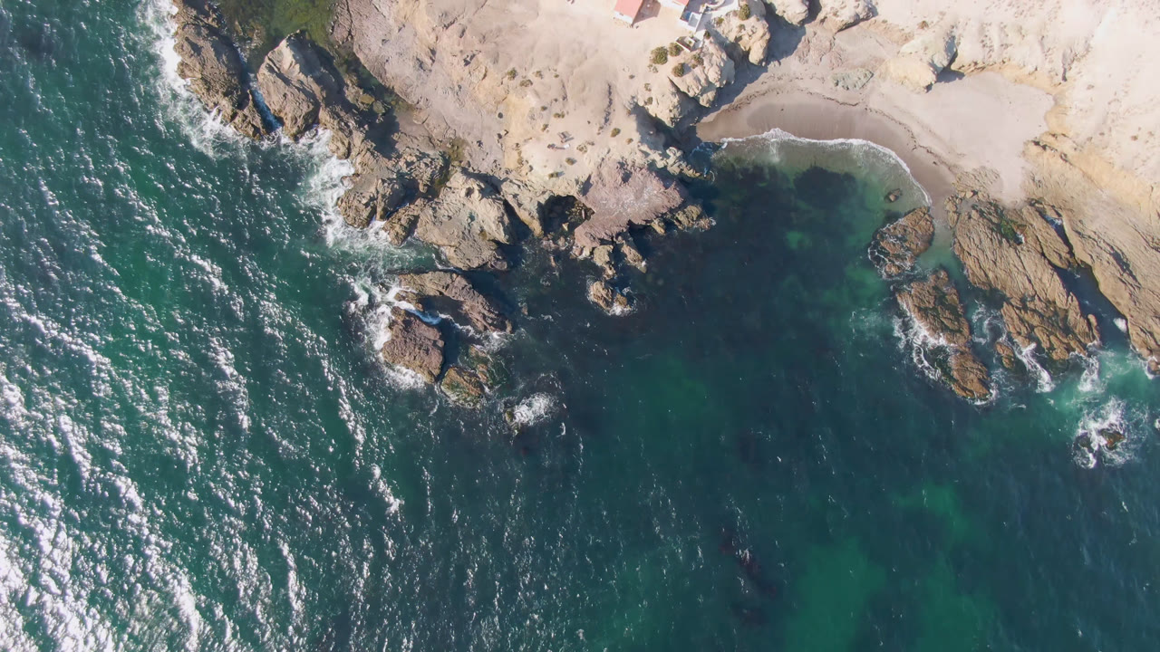Overhead view of rocky shoreline, teal water, sandy cove, and red-roofed buildings