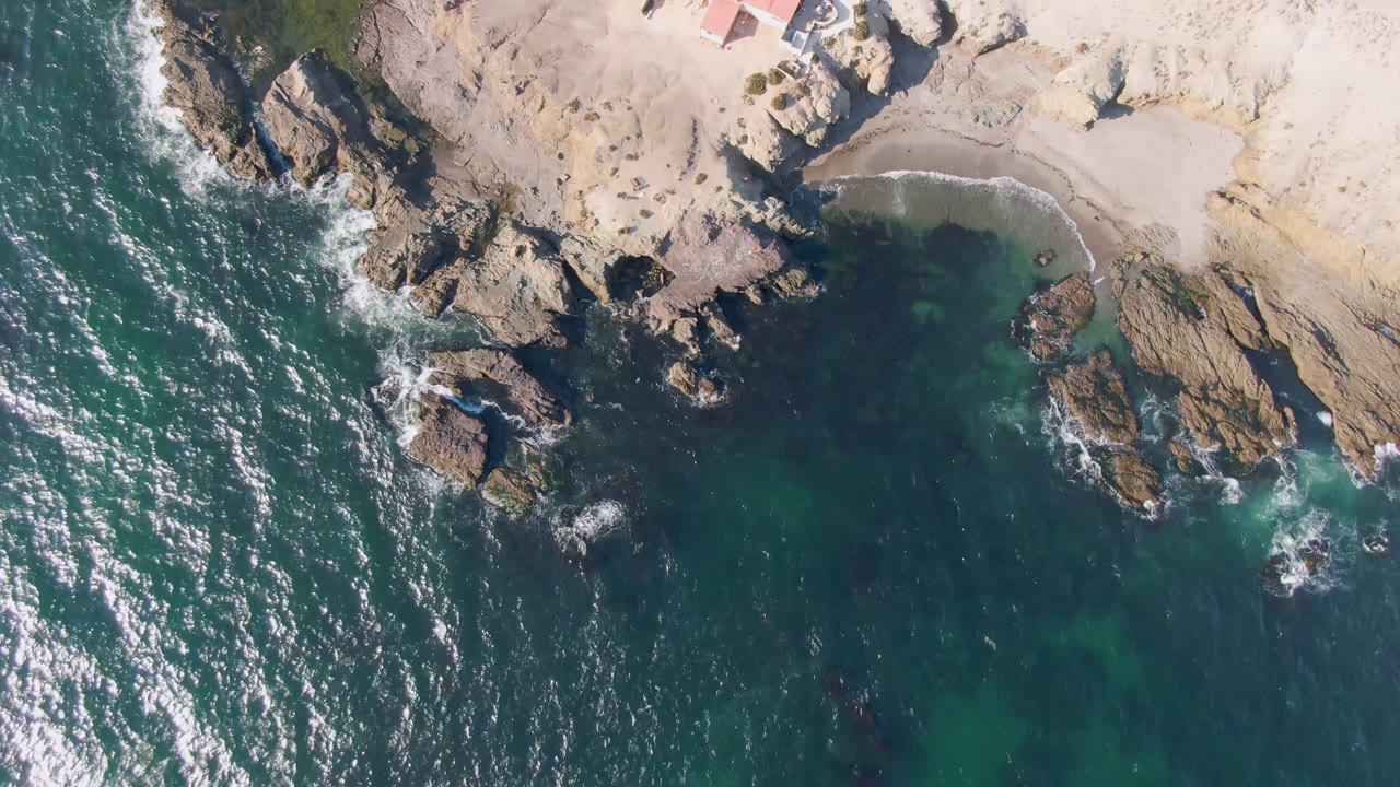 Bahia Ascuncion drone frame shows foamy surf along dark rocks, clear green-blue water, a curved sandy cove, and no visible people.