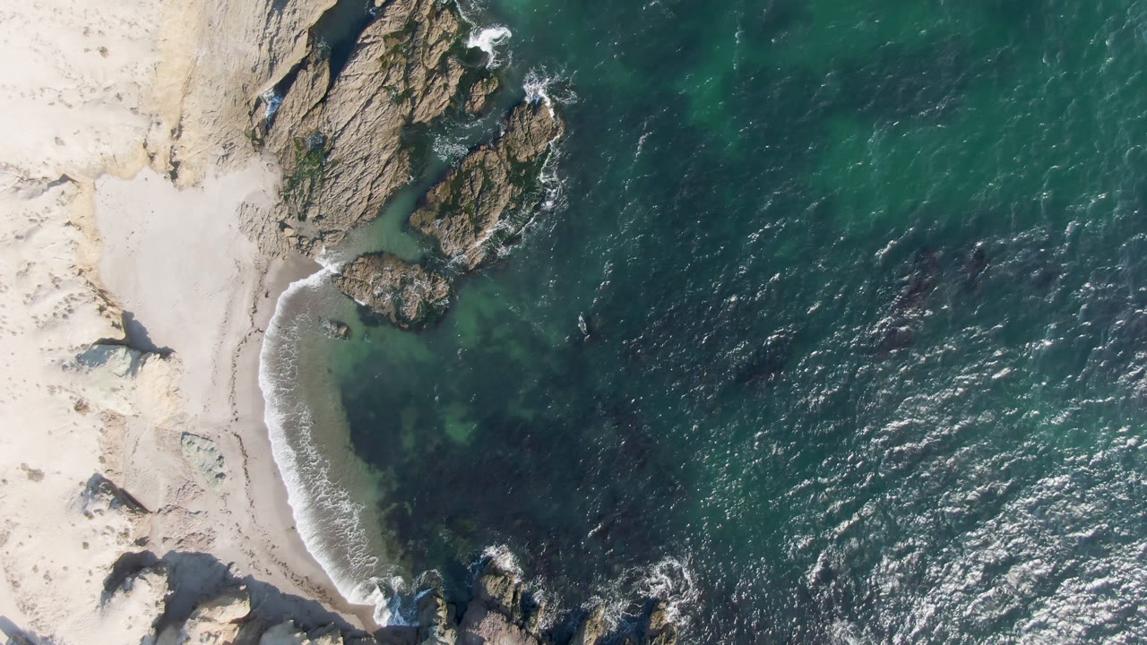 Overhead drone frame of Bahia Ascuncion showing pale sand at left, jagged rocks near the shoreline, green-blue ocean at right, and no visible people.