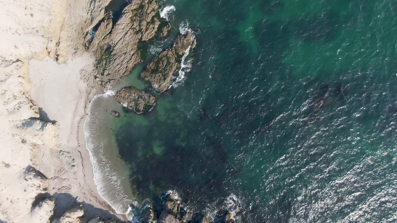 Aerial frame over Bahia Ascuncion with the rocky outcrop slightly more centered, surf curling along the sand, rippled ocean surface, and no visible people.