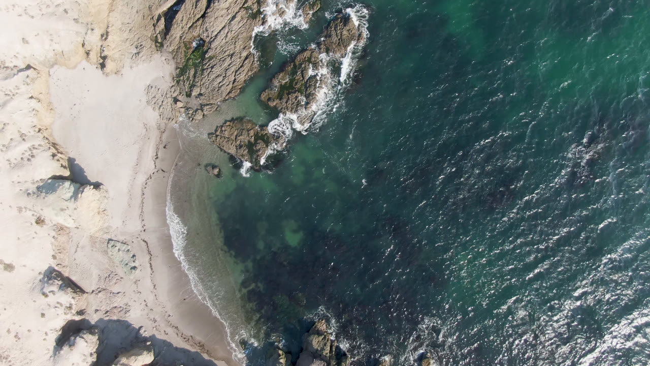Overhead frame of Bahia Ascuncion showing pale sand on the left, rocky outcrops along the shore, dark teal ocean to the right, and no visible people.