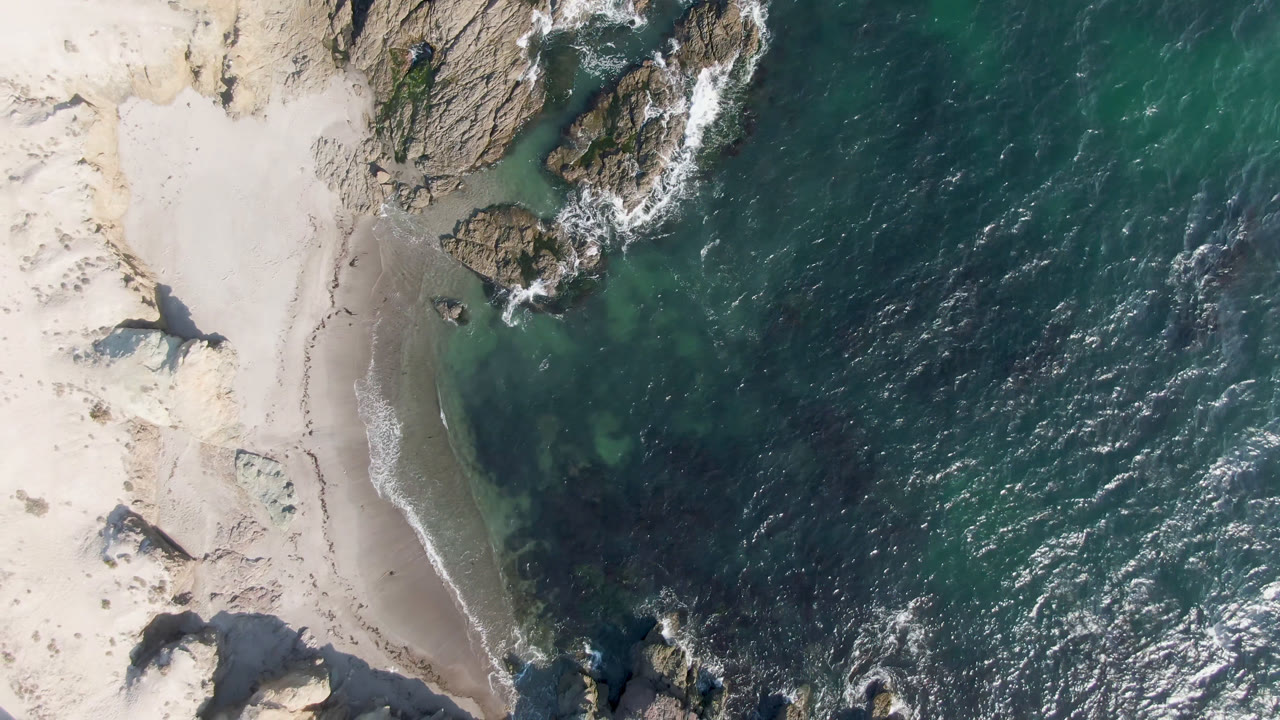Aerial frame over Bahia Ascuncion with a curved sand beach, a rocky point near the top, green shallows at the waterline, and no visible people.