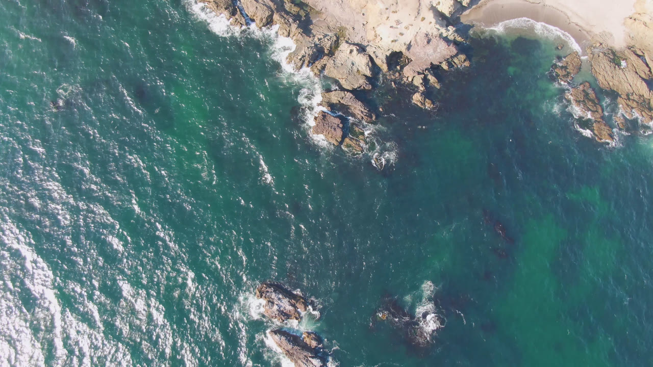 Overhead view of Bahia Ascuncion water with white foam, dark rocks near the upper shore, tan beach at the top right, and no visible people.