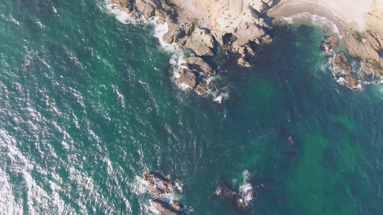 Aerial frame over Bahia Ascuncion showing sunlit water, foam streaks, offshore rocks near the lower center, pale sand along the top edge, and no visible people.