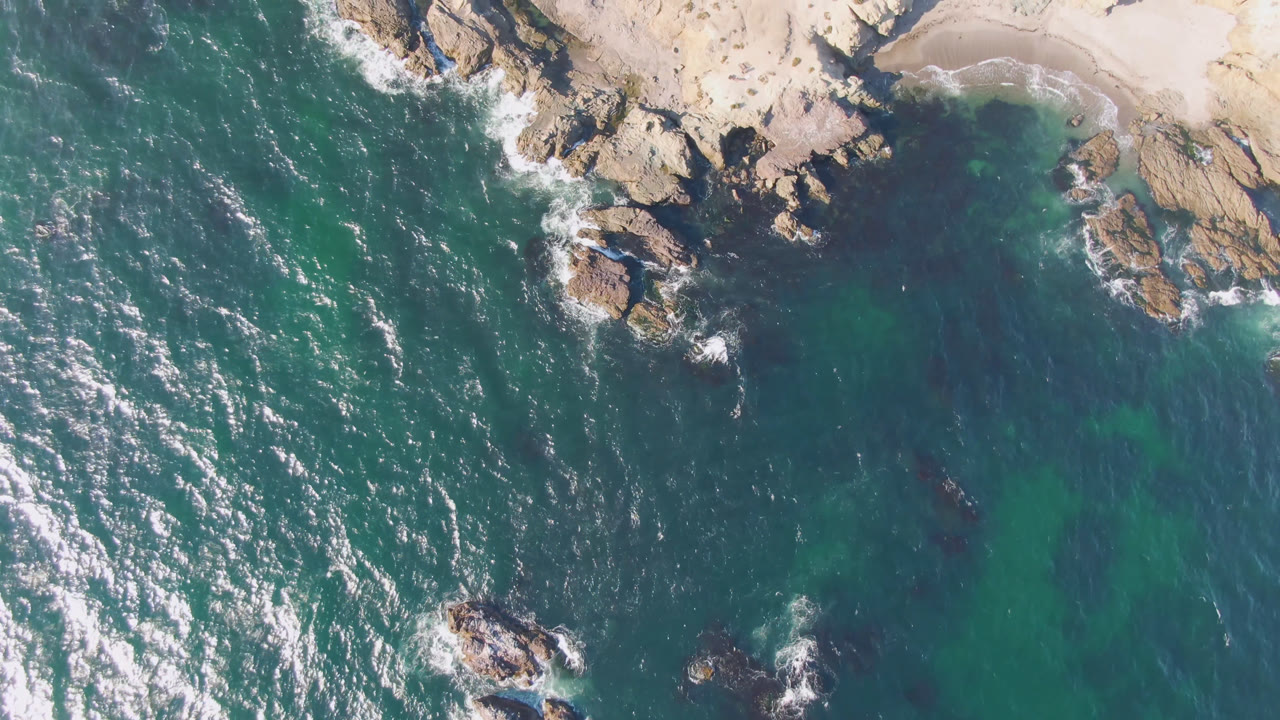 Top-down rocky cove with green-blue water and sand.