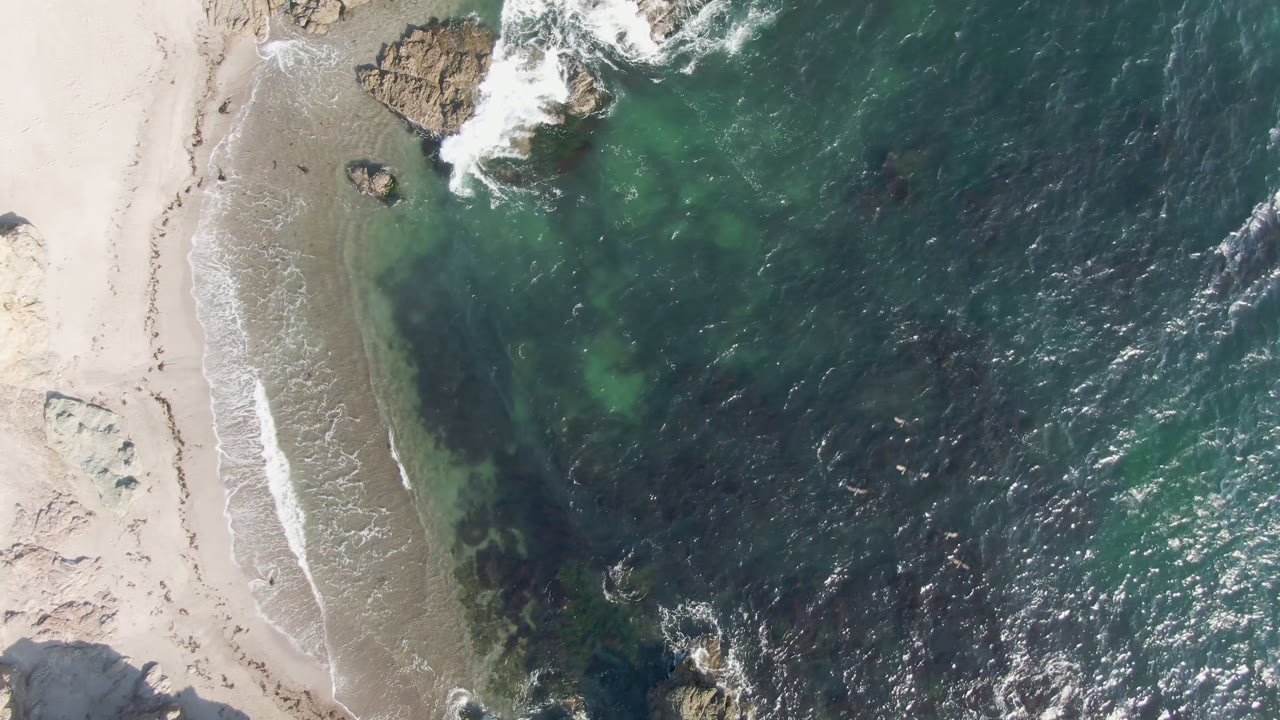 Overhead view of Bahia Ascuncion shoreline with pale sand on the left, green-blue shallows, dark ocean water, rocks near the top edge, and no visible people.