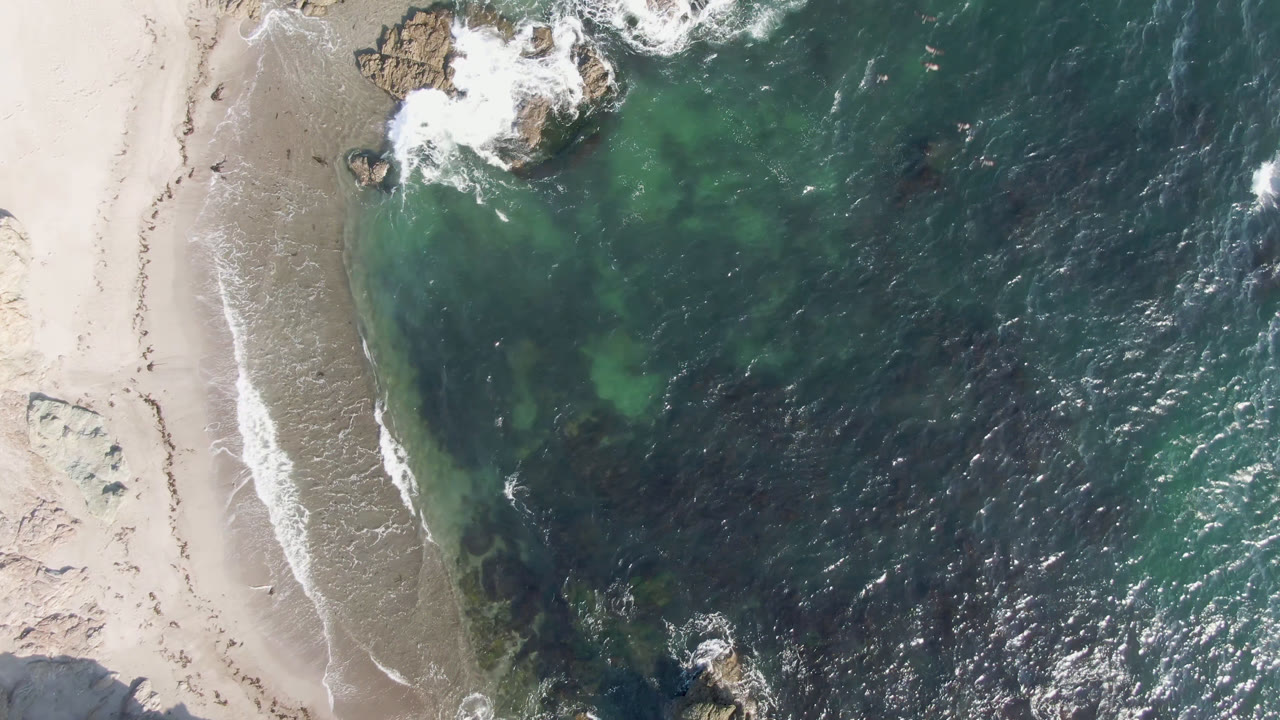 Aerial frame over Bahia Ascuncion showing a narrow sand strip, a fresh white wave line, rocky outcrops near the top and lower edge, and no visible people.