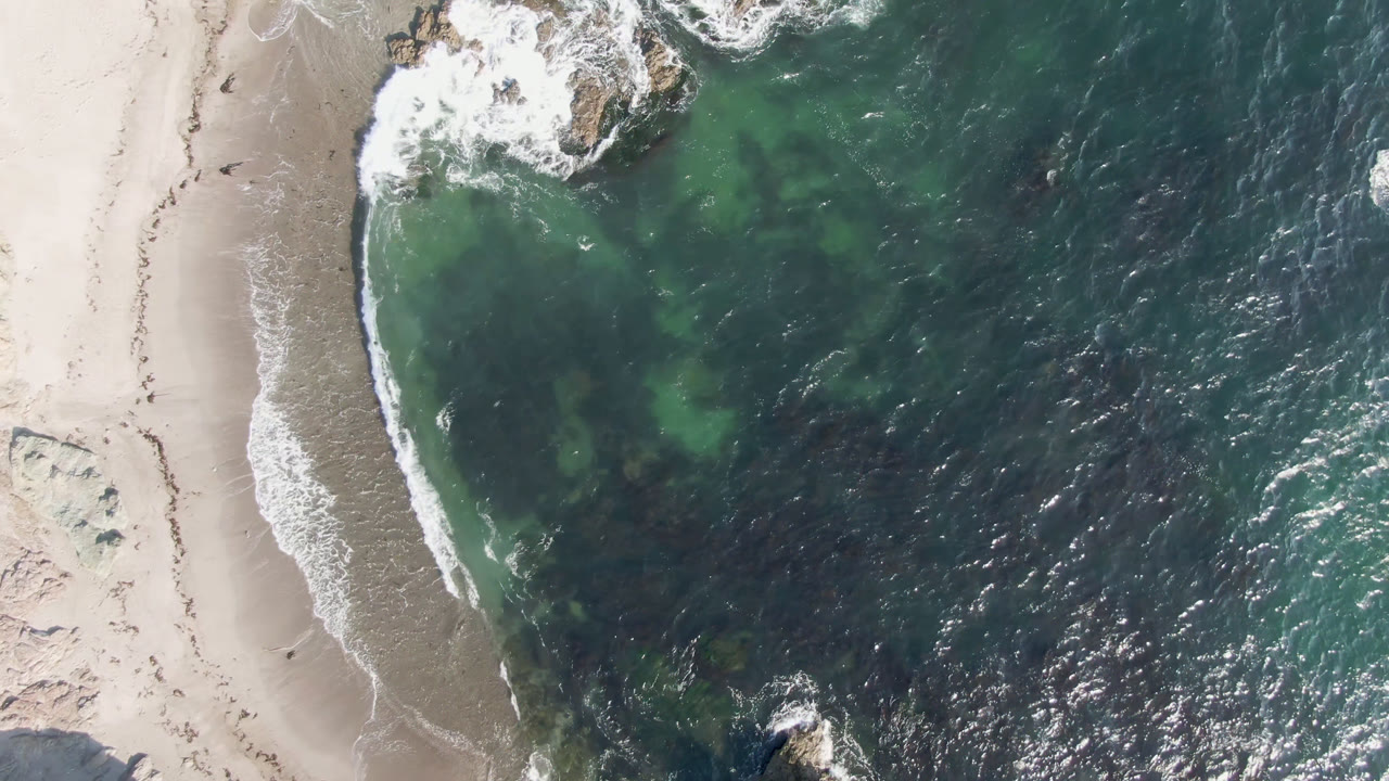 Top-down coast with foam around rocks, beach, and textured ocean.
