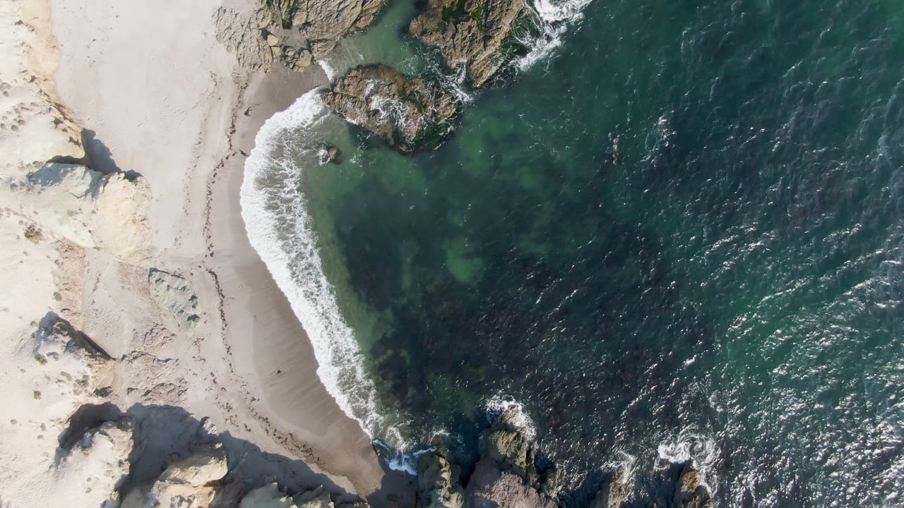 Overhead drone frame of Bahia Ascuncion beach with pale sand, rocky outcrops, green-blue water, white surf, and no visible people at the start of the clip.