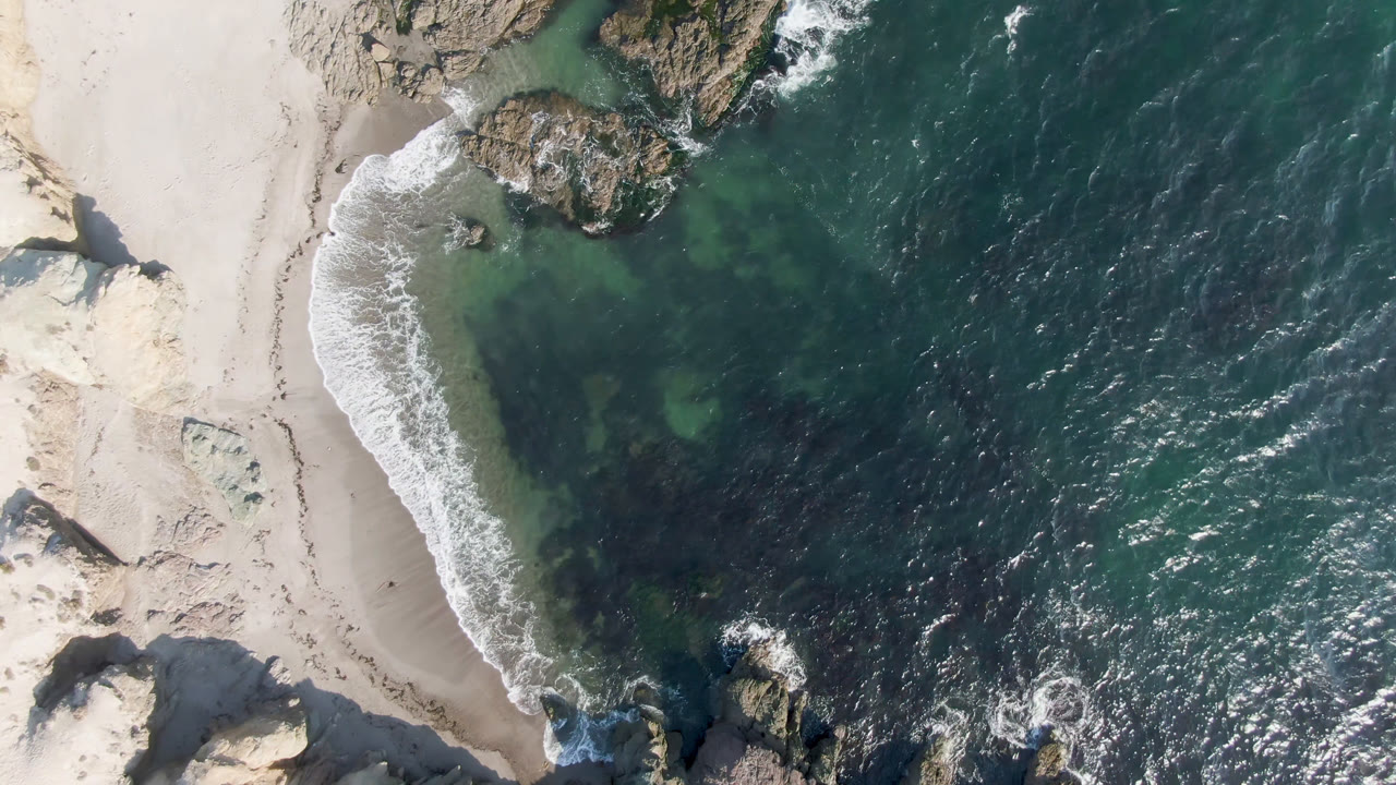 Aerial view over Bahia Ascuncion where a curved white surf line reaches the sand beside large rocks and darker offshore water, with no visible people.