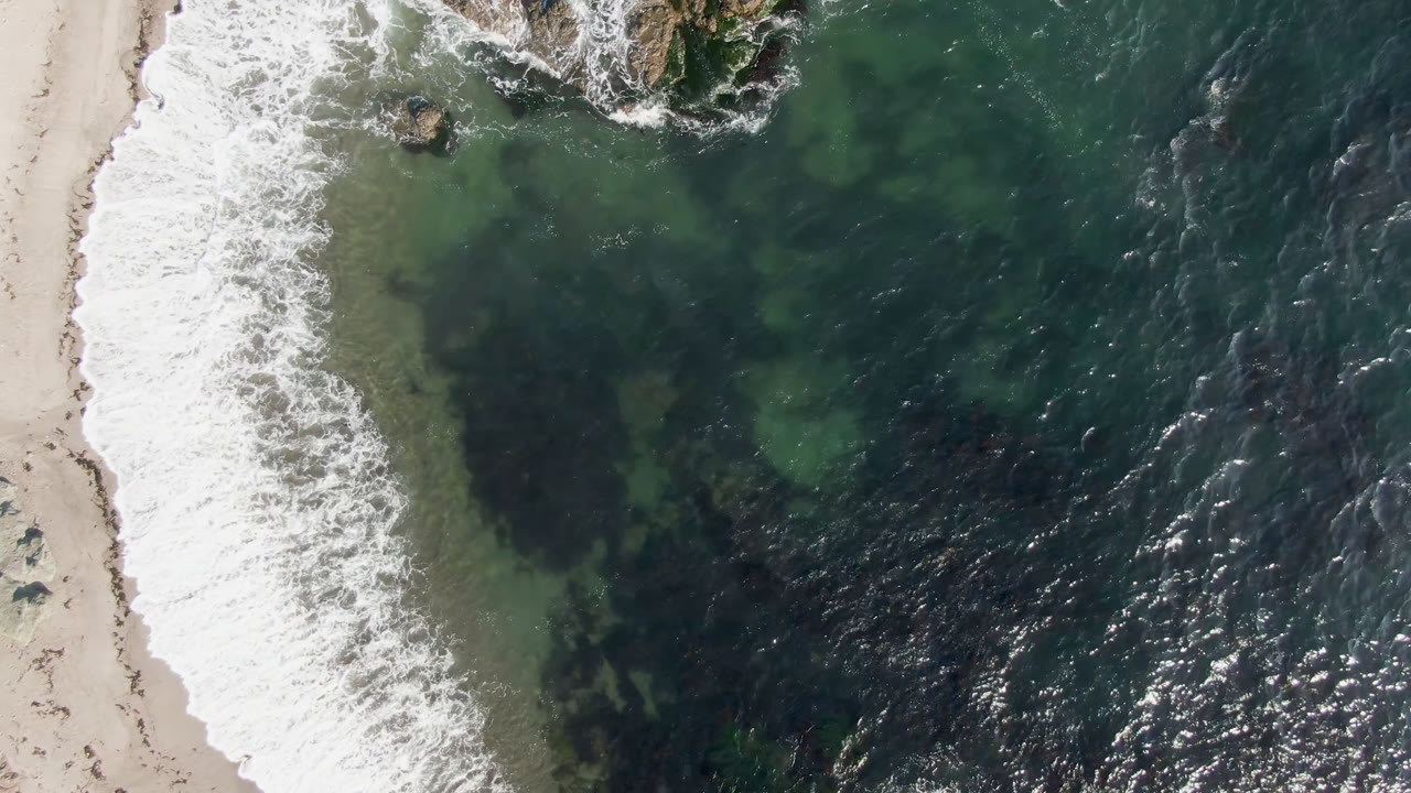 Top-down coast view with surf, sand, green water, and rocks.
