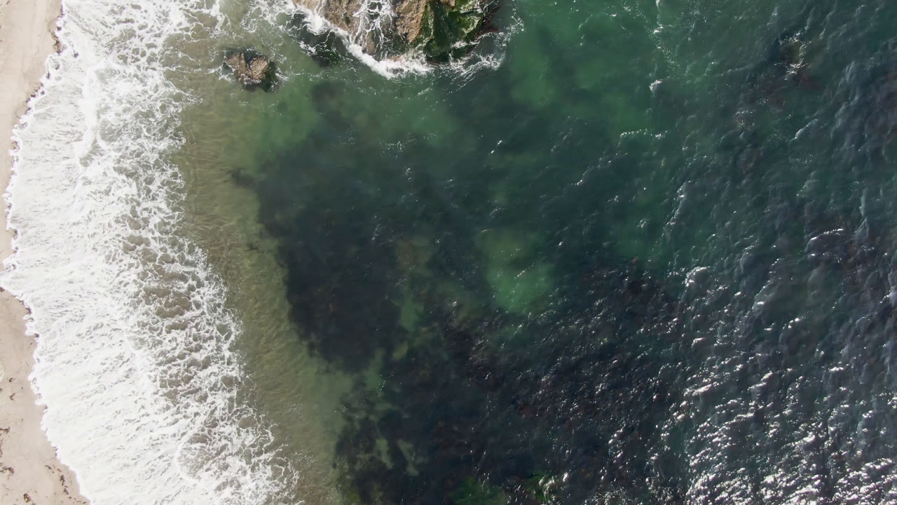 Aerial view over Bahia Ascuncion with a wide white wave band running along the sand, green nearshore water, dark reef-like patches below the surface, and no visible people.