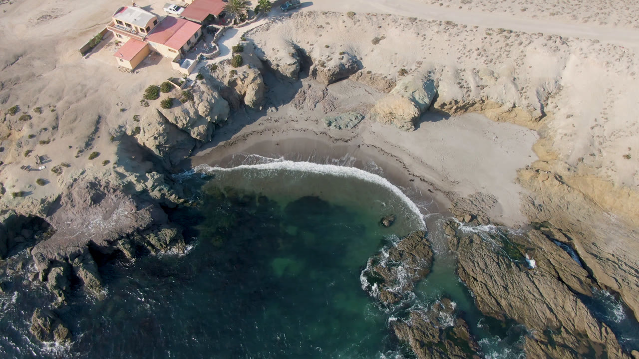 Aerial frame over Bahia Ascuncion showing dark green water, a curved strip of sand, rock outcrops, cliff edge, and red-roofed coastal buildings, with no visible people.
