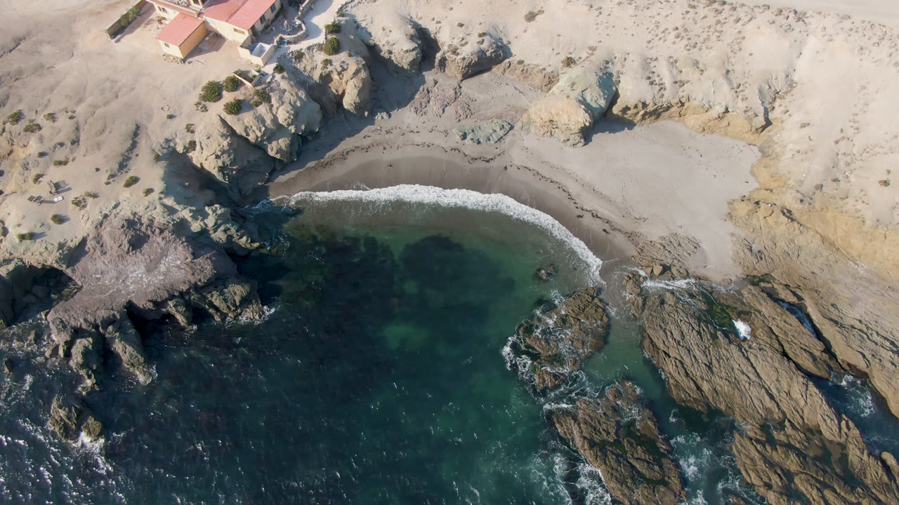Aerial view in Bahia Ascuncion with the sandy beach centered between pale cliffs and dark green ocean water, while a small building sits near the upper left edge, with no visible people.