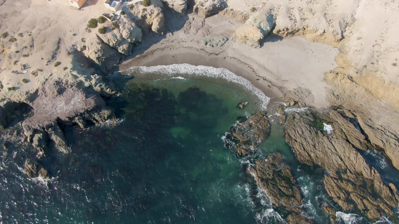 Overhead drone view of surf wrapping around rocks beside a sandy beach and cliff.