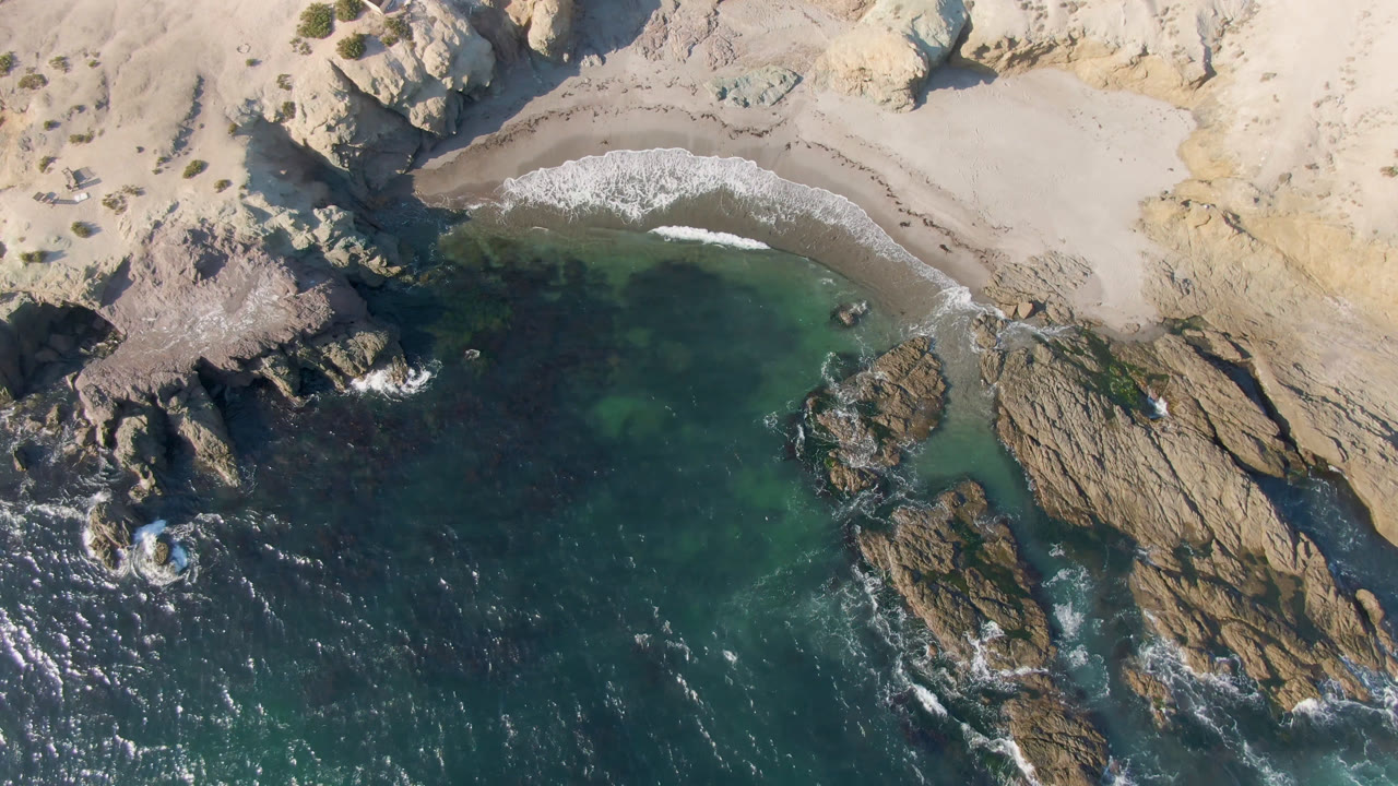Overhead frame from Bahia Ascuncion shows a pale sandy cove above dark green-blue water, with rocky outcrops at left and center and no visible people.