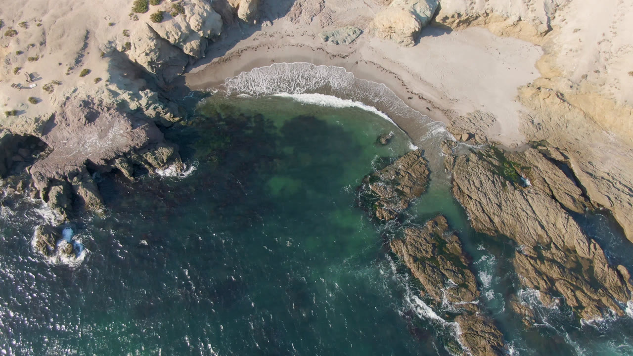 Aerial view in Bahia Ascuncion shows a white wave line reaching the small beach, dark water below, and tan rock formations framing the cove with no visible people.