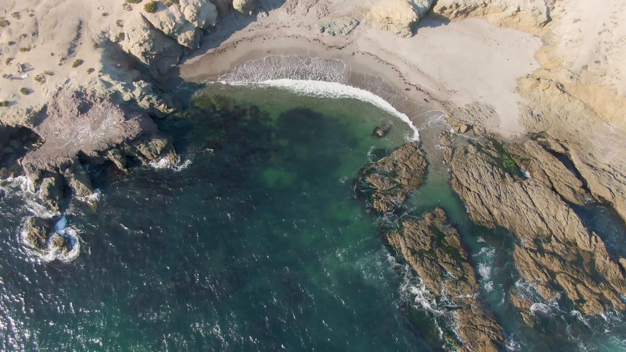 Drone view of dark ocean water, a sandy pocket beach, and jagged rocks.