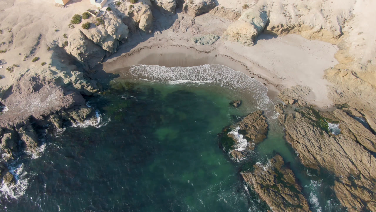 Overhead aerial frame of Bahia Ascuncion showing teal water, tan rock ledges, a pale sand cove, and a small house near the upper shore, with no visible people.