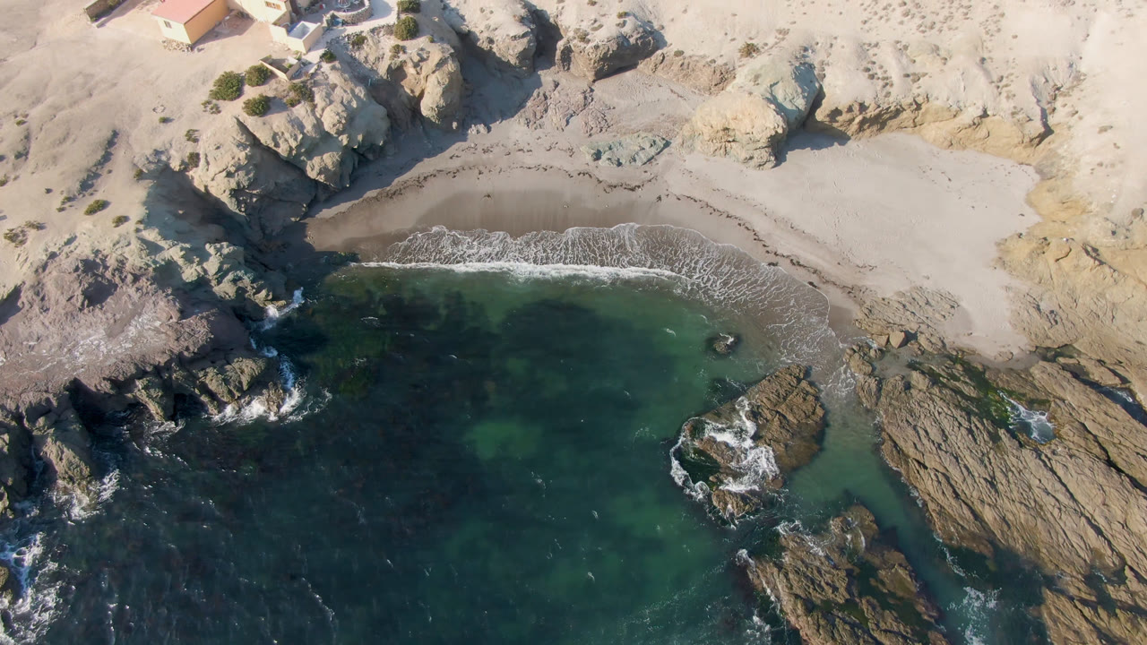 Aerial view over Bahia Ascuncion with dark green water below, a curved sand beach above, exposed rock shelves on both sides, and no visible people.