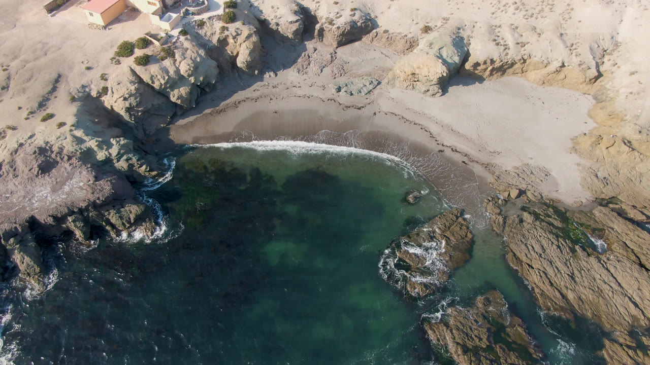 Rocky shoreline, pale beach, and shallow water seen from directly above.