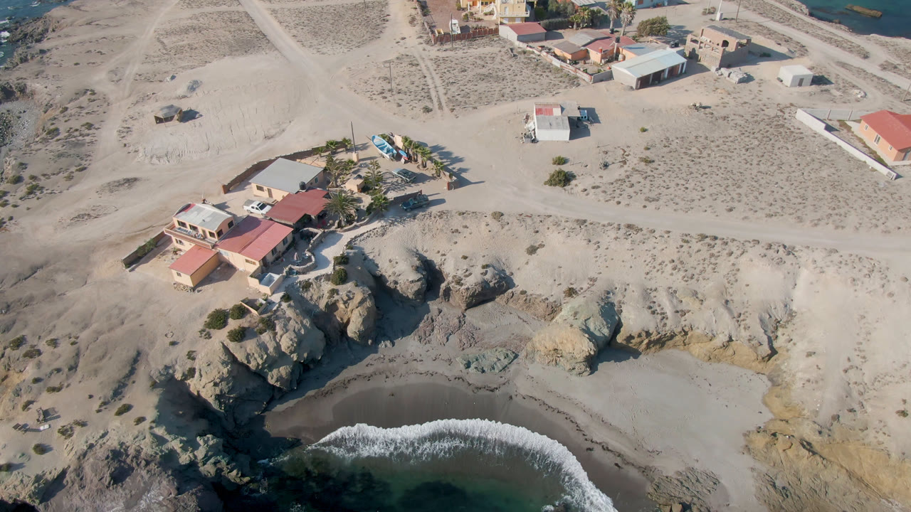 Aerial frame from Bahia Ascuncion with the red-roofed house near the top left, pale cliff across the middle, a wave curling onto the beach, and no visible people.