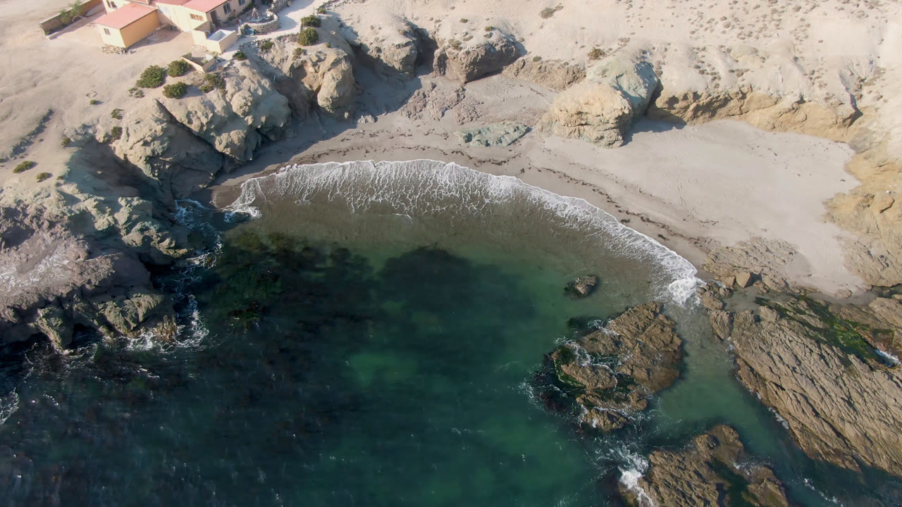 Aerial view of a rocky beach cove with green water and a house above the sand.