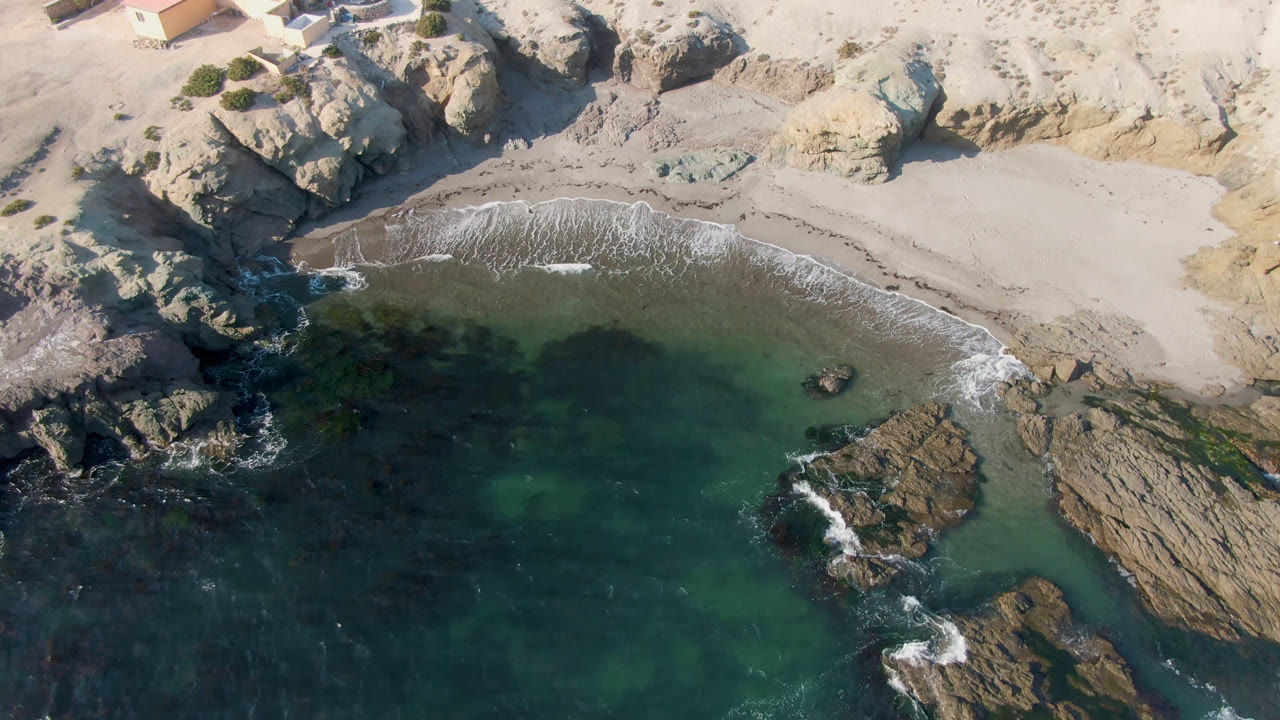 Aerial view of Bahia Ascuncion coastline with foamy surf reaching a curved sandy beach, dark rocks in the water, pale cliffs, and no visible people.
