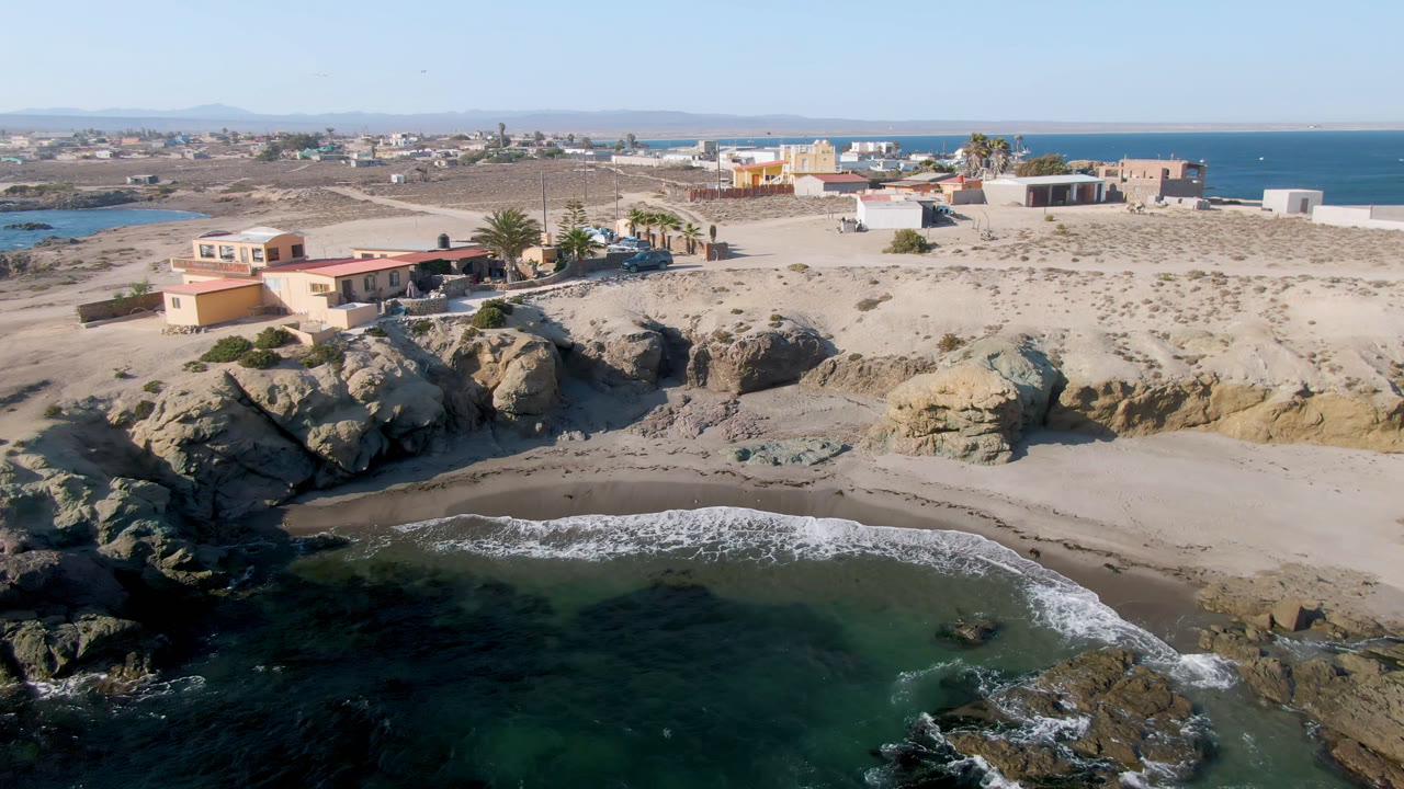 Aerial view of Bahia Ascuncion with the rocky point more centered, small waves on the beach, low coastal buildings beyond the sand, and no visible people.