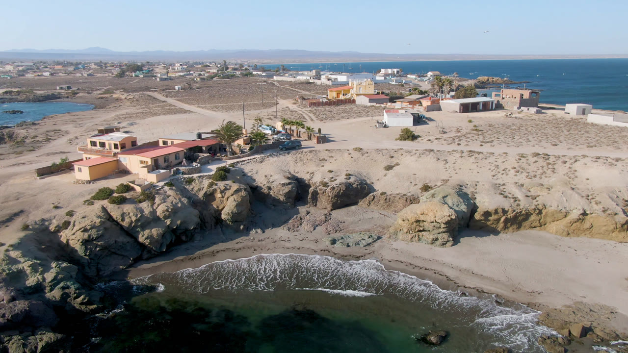 Aerial frame from Bahia Ascuncion showing dark green ocean at the bottom, a sandy cove below rocky bluffs, low buildings near the shore, open desert terrain, and no visible people.